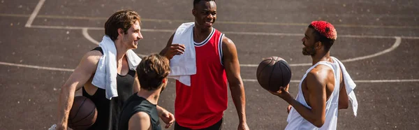 Positivo hombre afroamericano sosteniendo pelota de baloncesto cerca de amigos sonrientes con toallas al aire libre, pancarta - foto de stock