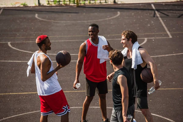 Uomo afroamericano sorridente mentre tiene palla da basket vicino agli amici con bottiglie d'acqua — Foto stock