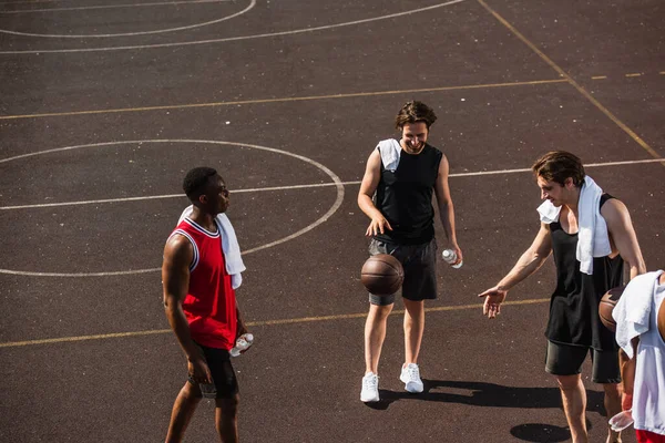 Улыбающийся мужчина играет в баскетбол рядом с межрасовыми друзьями с полотенцами и бутылками воды на детской площадке — стоковое фото