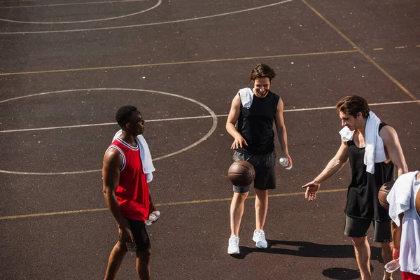 Smiling man playing basketball near interracial friends with towels and bottles of water on playground — Stock Photo