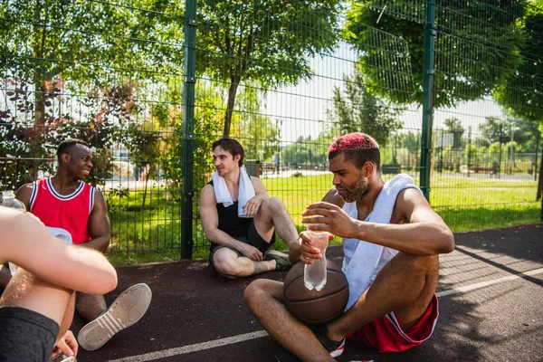 African american sportsman holding bottle of water near basketball ball and friends — Stock Photo
