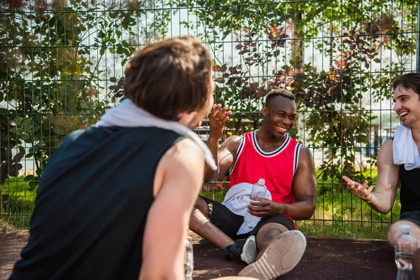 Cheerful african american sportsman with bottle of water talking to friends on playground — Stock Photo