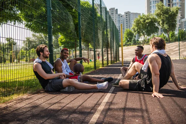Desportistas inter-raciais alegres com toalhas descansando no playground de basquete — Fotografia de Stock