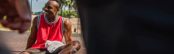 Positive african american sportsman with towel sitting near water on playground, banner — Stock Photo