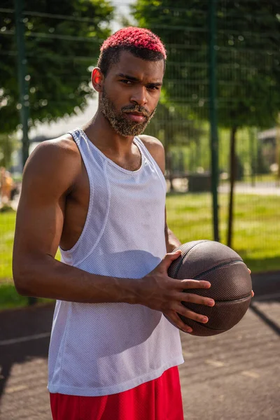 African american sportsman with dyed hair holding basketball ball and looking at camera outdoors — Stock Photo