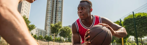 Enfocado afroamericano deportista sosteniendo pelota de baloncesto cerca borrosa jugador, bandera - foto de stock