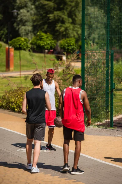Улыбающийся африканский американский спортсмен стоит рядом с друзьями и баскетбольный мяч на открытом воздухе — стоковое фото