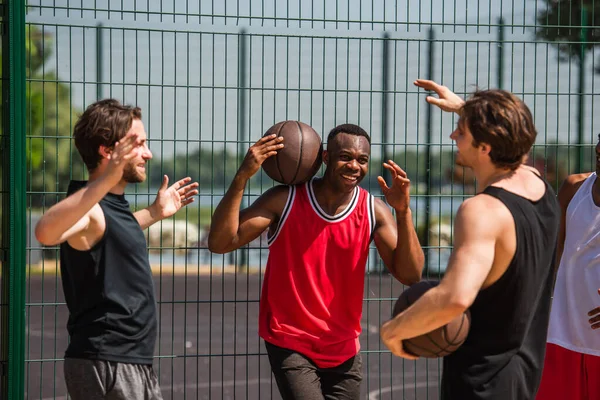 Amis multiethniques positifs en vêtements de sport tenant des balles de basket près de la clôture à l'extérieur — Photo de stock