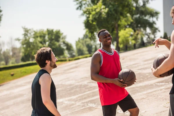 Alegre jugador de baloncesto afroamericano con pelota mirando a amigos al aire libre - foto de stock