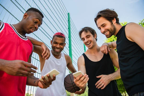 Vista de ángulo bajo de jugadores de baloncesto interracial positivos usando teléfonos inteligentes - foto de stock