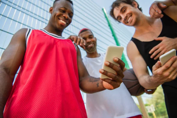 Vista a basso angolo dello smartphone in mano di uno sportivo africano americano sorridente vicino agli amici su sfondo sfocato — Foto stock