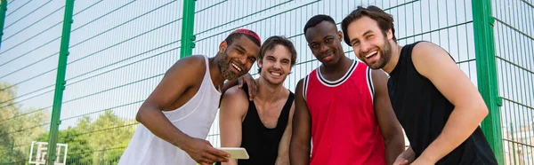 Deportistas interraciales con smartphone sonriendo a la cámara al aire libre, pancarta — Stock Photo