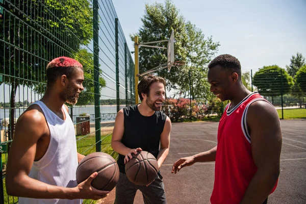 Uomo sorridente che tiene la pallacanestro vicino agli amici americani africani sul parco giochi — Foto stock