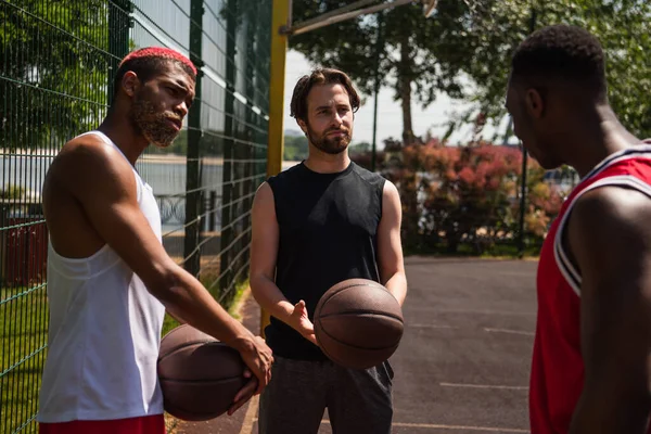 Jóvenes deportistas con pelotas de baloncesto mirando a un amigo afroamericano en el patio de recreo - foto de stock