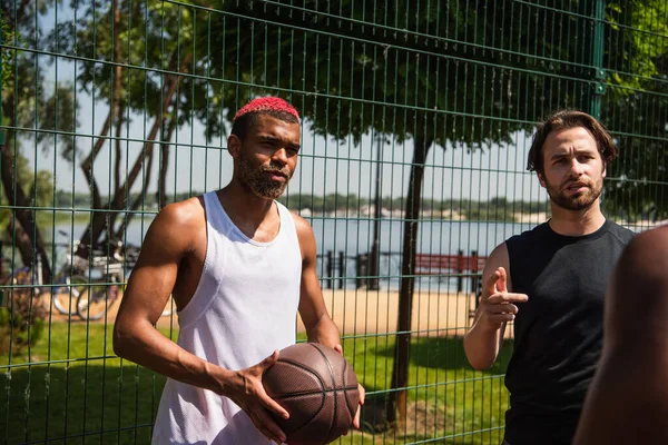 Jugador de baloncesto afroamericano de pie cerca de amigos hablando al aire libre - foto de stock