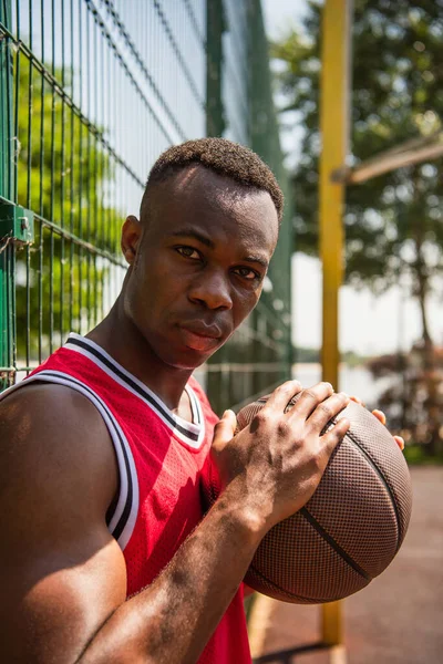 African american sportsman with basketball ball looking at camera near fence — Stock Photo