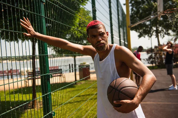 Africano americano desportista com cabelo tingido segurando bola de basquete perto de cerca na quadra — Fotografia de Stock