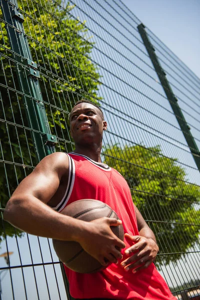 Vista de ángulo bajo del sonriente deportista afroamericano sosteniendo pelota de baloncesto cerca de la valla - foto de stock