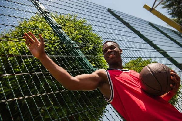 Low angle view of positive african american sportsman holding basketball ball and waving hand near fence — Stock Photo