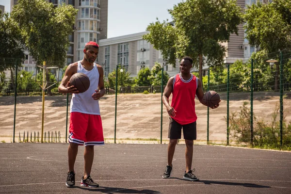 Smiling african american sportsman looking at friend with basketball ball on court — Stock Photo