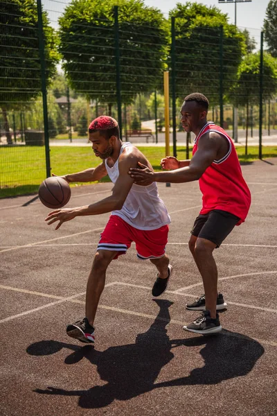 Vista lateral del deportista afroamericano con pelota de baloncesto corriendo en un parque infantil cerca de un amigo - foto de stock