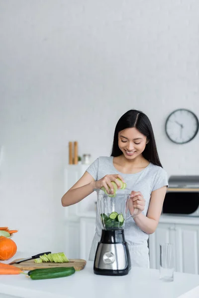 Happy asian woman adding sliced cucumbers into shaker near fresh vegetables and chopping board — Stock Photo