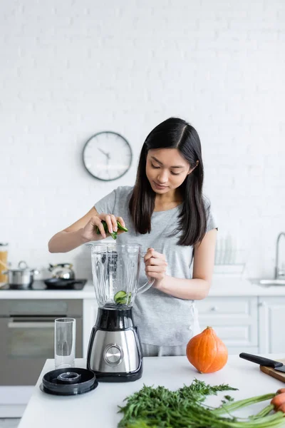 Junge asiatische Frau fügt geschnittene Gurke in elektrischen Shaker während der Zubereitung des Frühstücks — Stockfoto