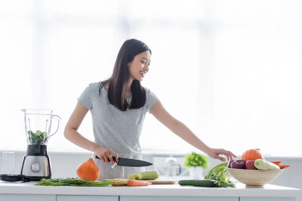 Feliz mulher asiática com faca preparando café da manhã de legumes frescos na cozinha — Fotografia de Stock