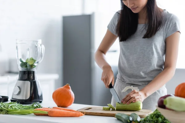 Vue partielle de femme coupant des courgettes près du mélangeur, de la citrouille crue et des carottes sur la table — Photo de stock