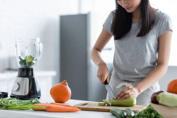 Vista parcial de la mujer cortando calabacín cerca de licuadora, calabaza cruda y zanahorias en la mesa - foto de stock