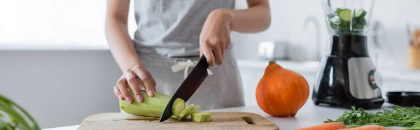 Partial view of woman cutting zucchini near ripe pumpkin and blender with sliced cucumber, banner — Stock Photo