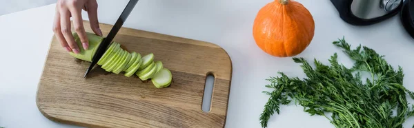 Vista parziale della donna che taglia zucchine fresche sul tagliere vicino alla zucca matura sul tavolo, striscione — Foto stock