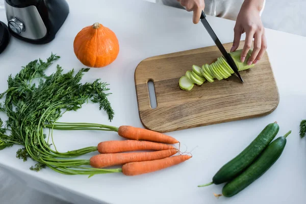 Vista parcial de la mujer cortando calabacín en la tabla de cortar cerca de verduras frescas en la cocina - foto de stock