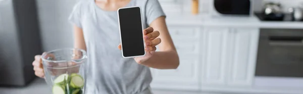 Cropped view of blurred woman showing smartphone with blank screen near shaker with zucchini, banner — Stock Photo
