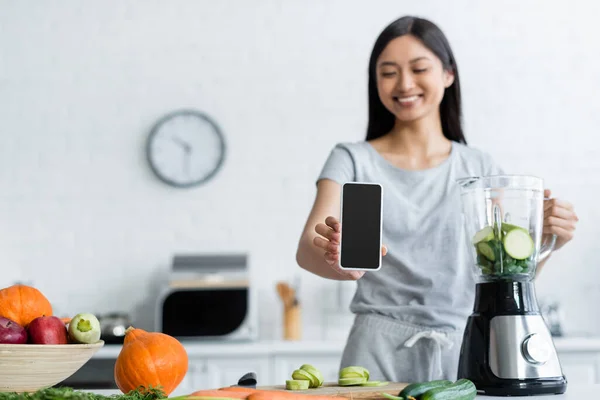 Donna asiatica offuscata mostrando cellulare con schermo vuoto vicino frullatore e verdure fresche — Foto stock