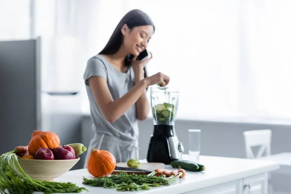 Desenfocado asiático mujer hablando en celular cerca de fresco verduras y eléctrico agitador en cocina - foto de stock