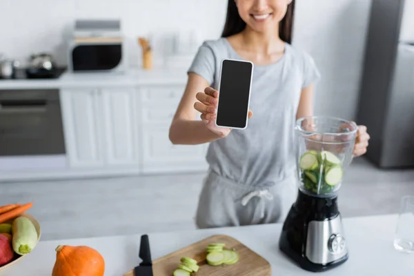 Vista ritagliata di donna offuscata che mostra smartphone con schermo bianco vicino frullatore e verdure fresche — Foto stock