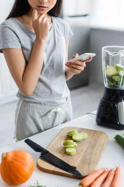 Vista recortada de la mujer en busca de receta en el teléfono inteligente cerca de verduras frescas y coctelera - foto de stock