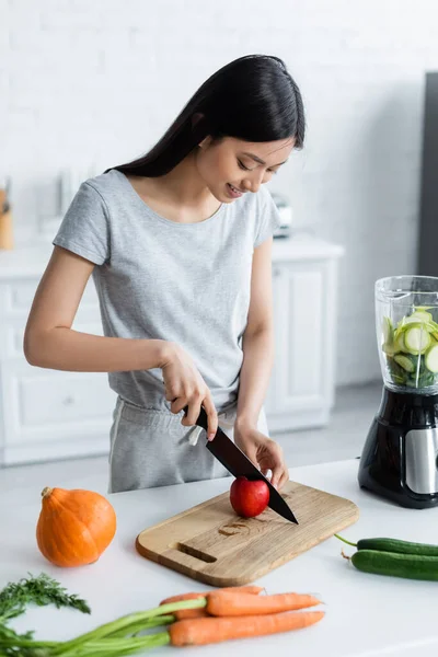 Позитивная азиатка режет яблоко рядом с электрическим блендером и овощами на кухонном столе — стоковое фото