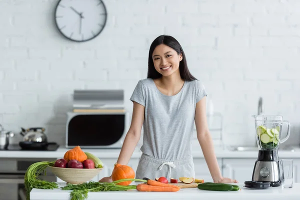 Allegra donna asiatica guardando la fotocamera vicino frullatore elettrico e verdure fresche in cucina — Foto stock