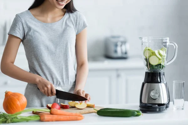 Cropped view of happy asian woman cutting apple near electric blender and fresh vegetables on kitchen table — Stock Photo