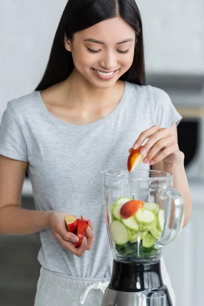 Smiling asian woman adding cut apple into electric shaker with fresh zucchini — Stock Photo