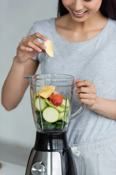 Partial view of smiling asian woman adding apple and zucchini into electric blender — Stock Photo