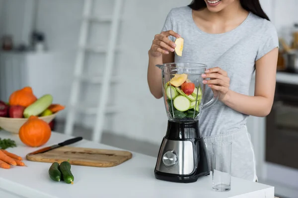 Partial view of smiling woman adding apple into electric blender with sliced zucchini — Stock Photo
