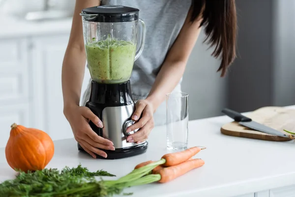 Vista parziale della donna che prepara frullato fresco vicino a zucca e carote sul tavolo della cucina — Foto stock