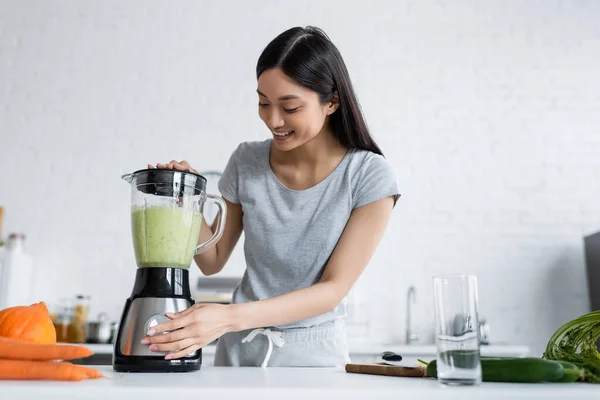 Cheerful asian woman preparing vegetable smoothie in electric shaker — Stock Photo