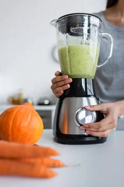 Vista recortada de la mujer preparando batido de verduras cerca de calabaza y zanahorias borrosas - foto de stock