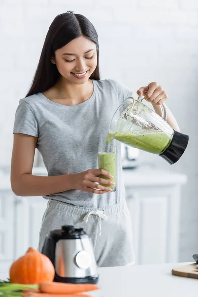 Alegre ásia mulher derramando caseiro smoothie no vidro perto borrado legumes no cozinha — Fotografia de Stock