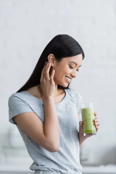 Joyful asian woman fixing hair while holding glass of fresh smoothie — Stock Photo