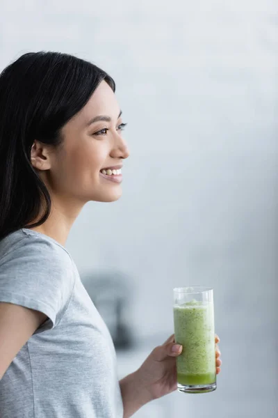 Giovane donna asiatica sorridente mentre tiene in mano un bicchiere di frullato fresco a casa — Foto stock